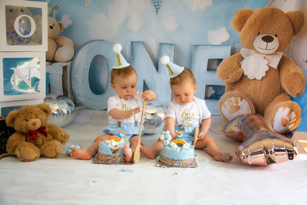 Twin babies celebrate their first birthday with matching cakes and teddy bear decorations.