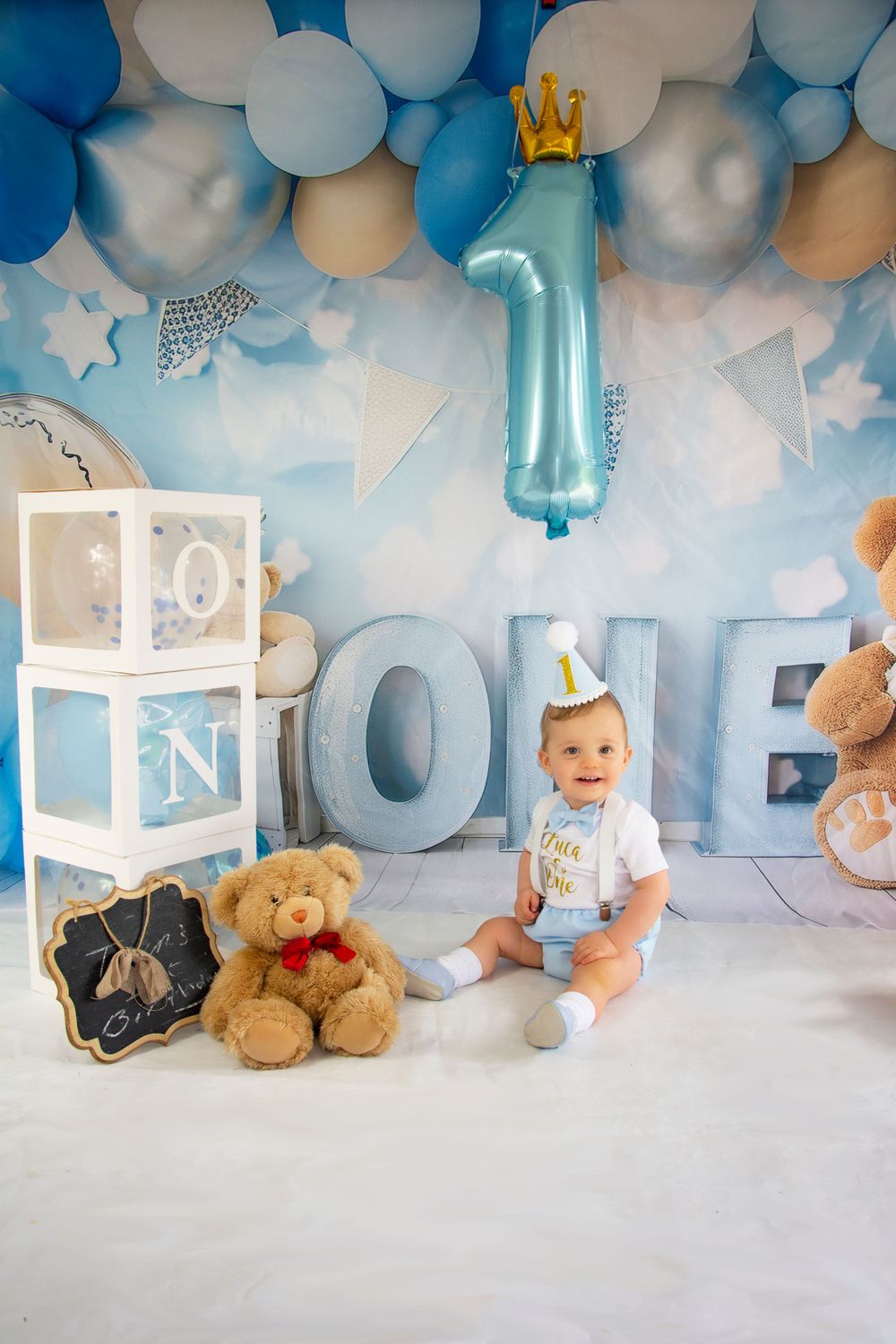 First birthday photoshoot with blue balloon decor, crown, and baby sitting beside ONE letters.