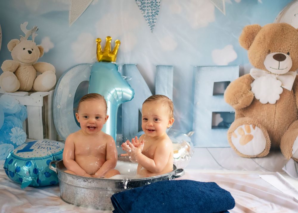 Twin babies sitting on blue fabric with crown decor and teddy bears for first birthday photoshoot.