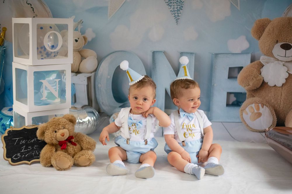 Twin babies in matching outfits sitting beside decorative boxes and teddy bears.