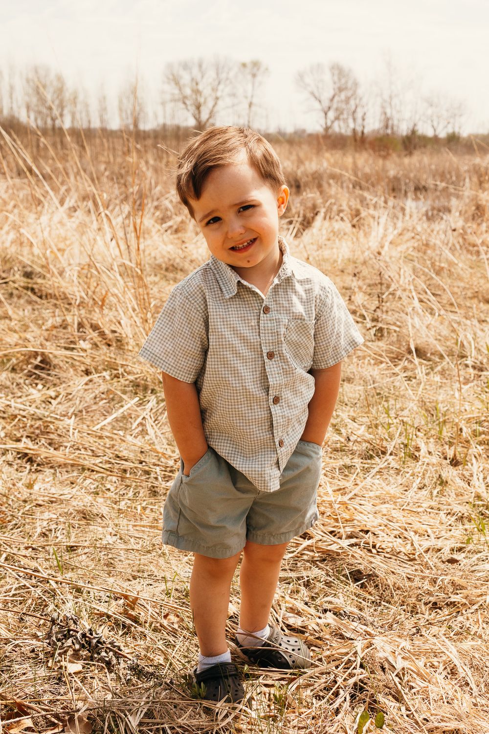 Smiling toddler boy in plaid shirt and green shorts standing in a golden field on a sunny day.