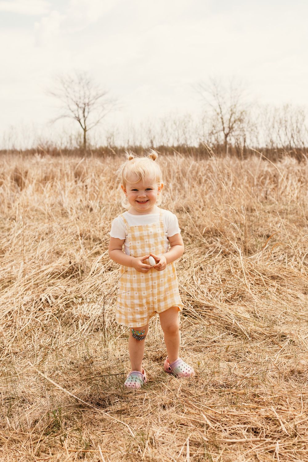 A toddler girl in a yellow gingham romper smiles while standing in a dry grassy field.