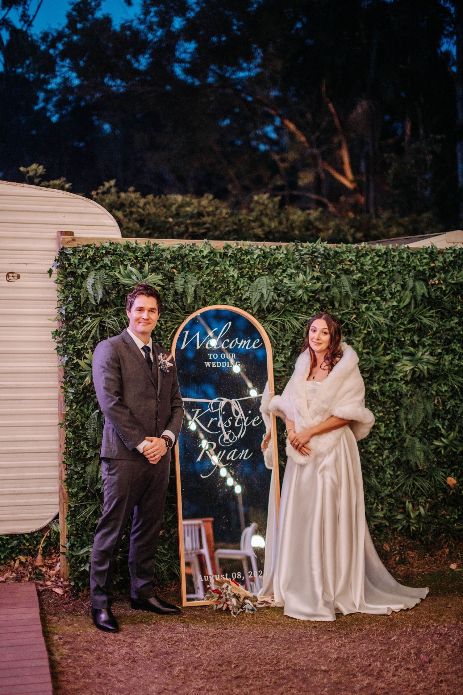 Vintage wedding welcome sign displayed between newlyweds next to white caravan trailer in outdoor garden setting.