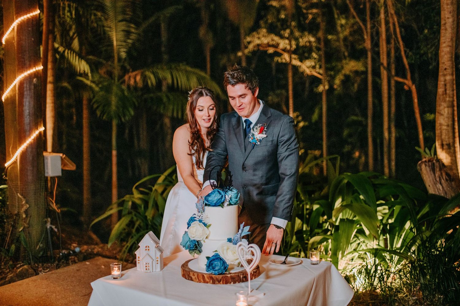 Wedding couple cuts elegant white and blue tiered cake at outdoor evening reception.