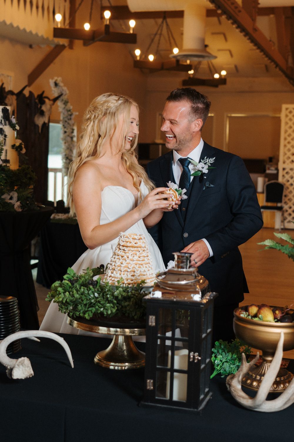 Couple laughs while serving cake at reception with rustic decor and antler details.