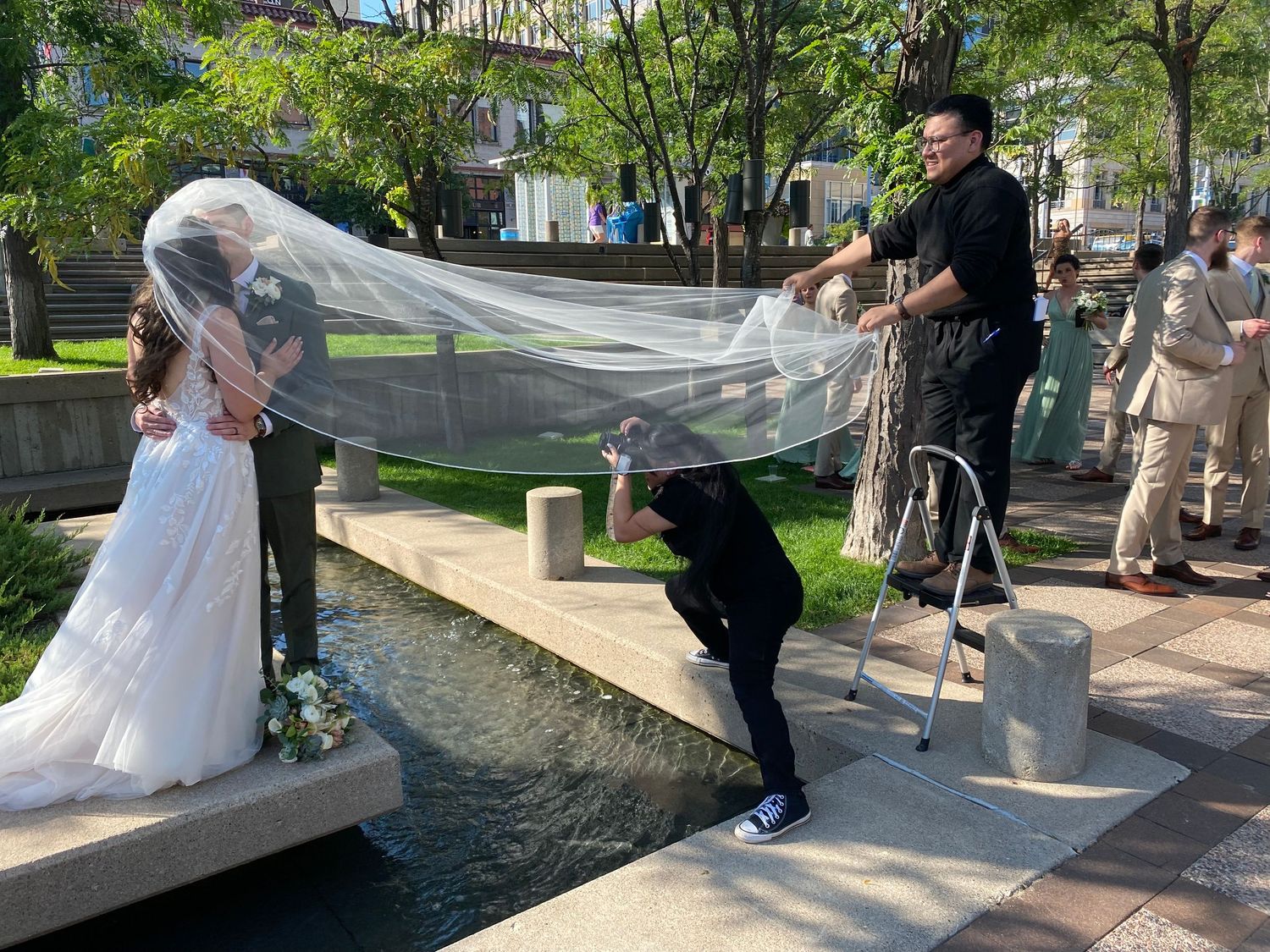 A photographer captures a wedding moment by a fountain with flowing veil in the breeze.
