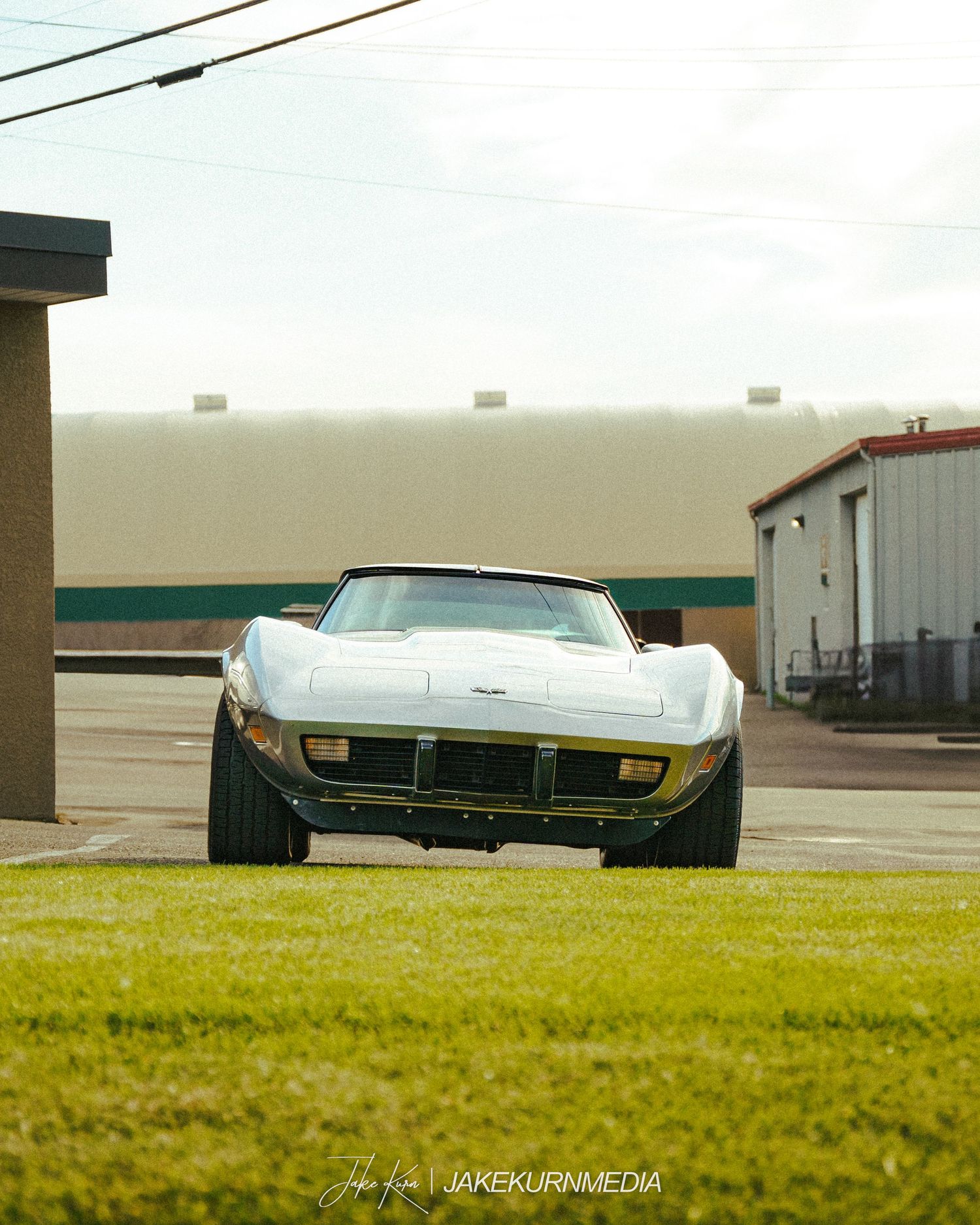 A vintage silver Chevrolet Corvette Stingray on grass with buildings in the background.