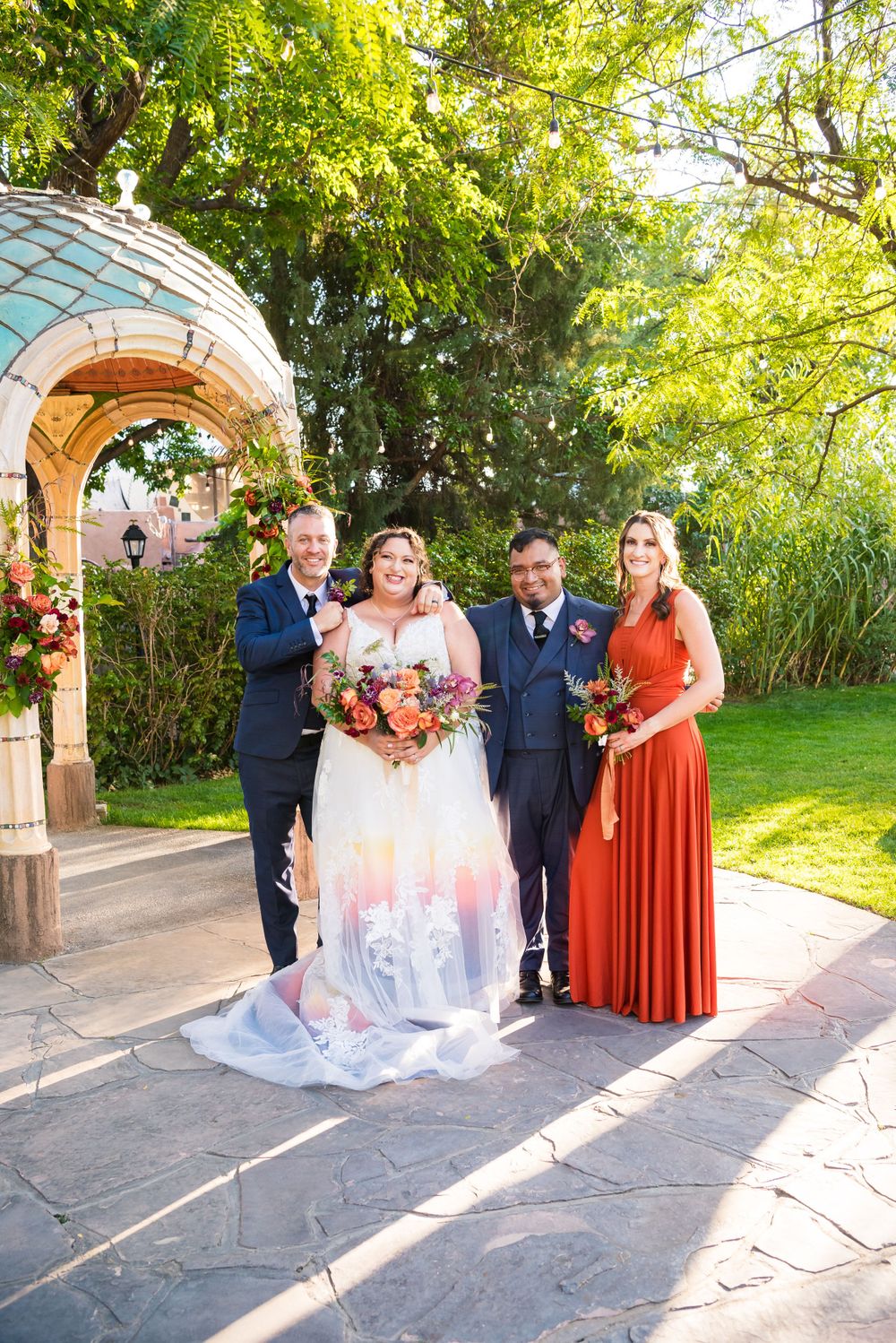 Wedding group photo under an ornate turquoise gazebo in a garden setting.