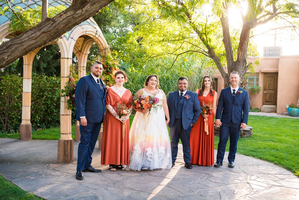 Wedding party in navy suits and coral dresses poses under a garden archway at sunset.