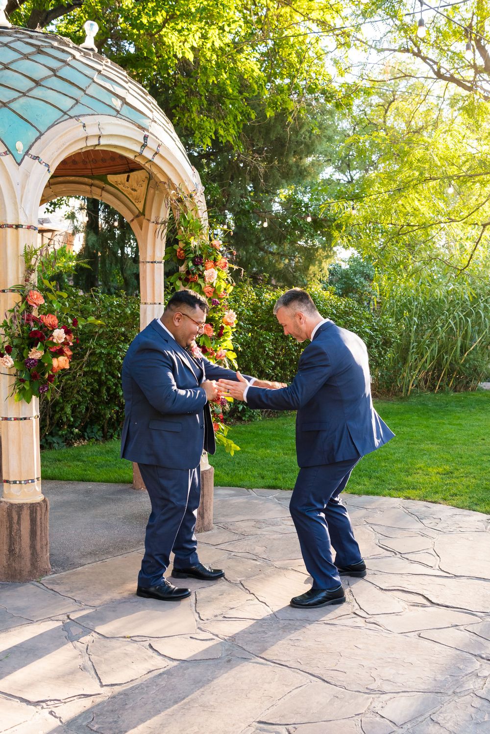 Two groomsmen in navy suits share a playful moment under a decorated garden archway.