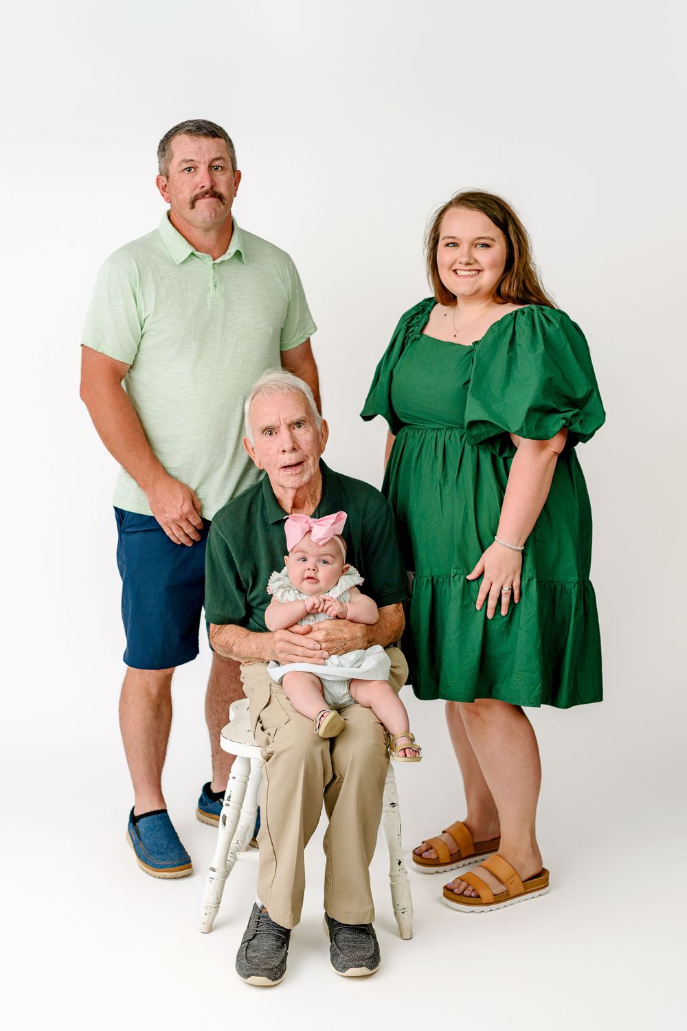 Multi-generational family portrait including three adults and a baby against white backdrop, showing casual attire.