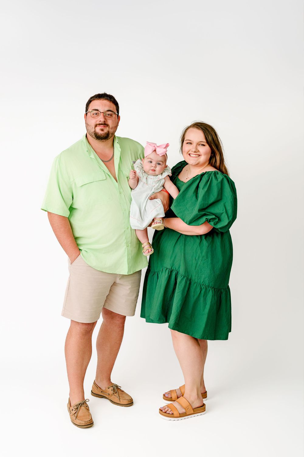 A family in coordinating green and neutral outfits poses together against a white studio backdrop.