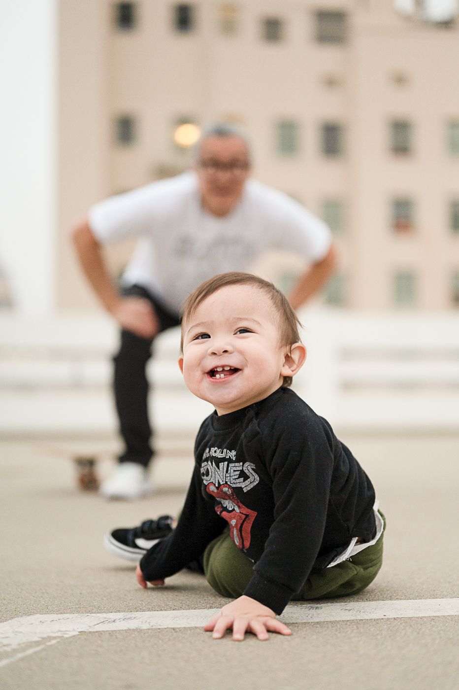 Joyful baby in dark clothing crawls on rooftop with blurred figure in background.