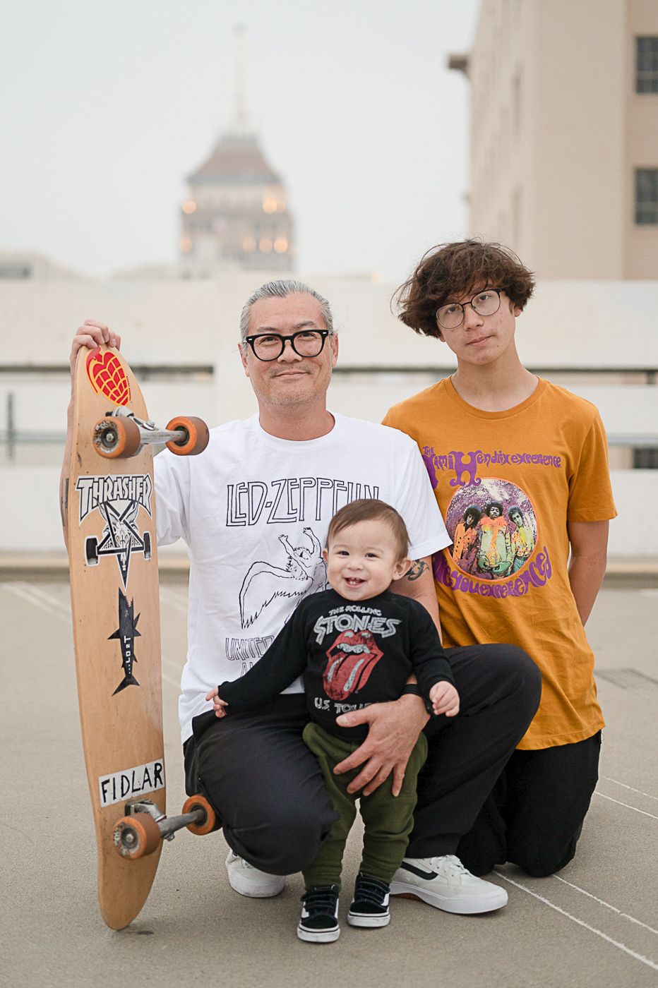 Three people pose together with decorated skateboard on urban rooftop.