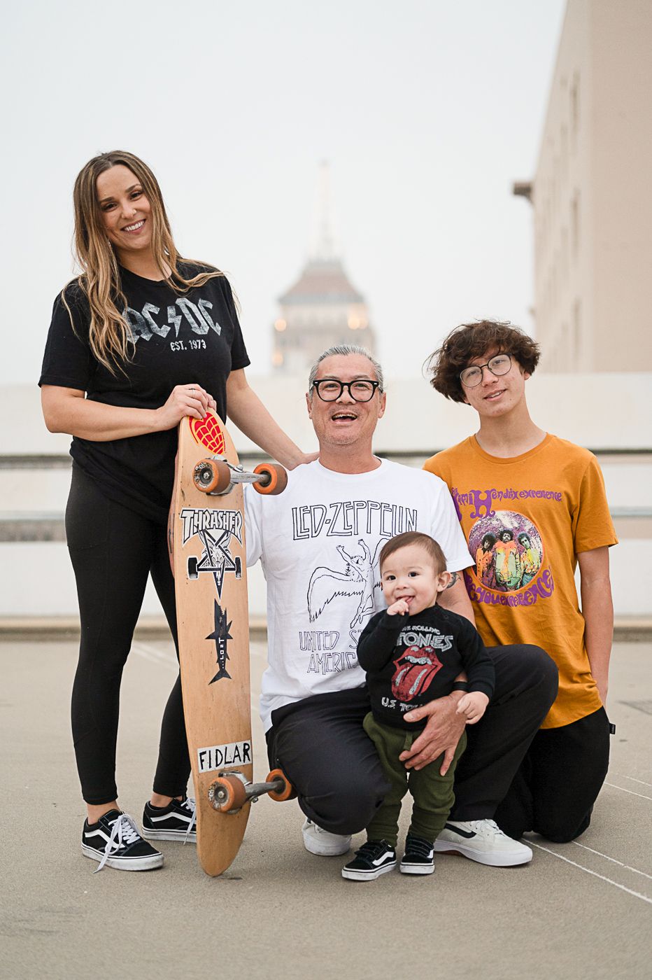 Family group poses with decorated skateboard on rooftop against city backdrop.