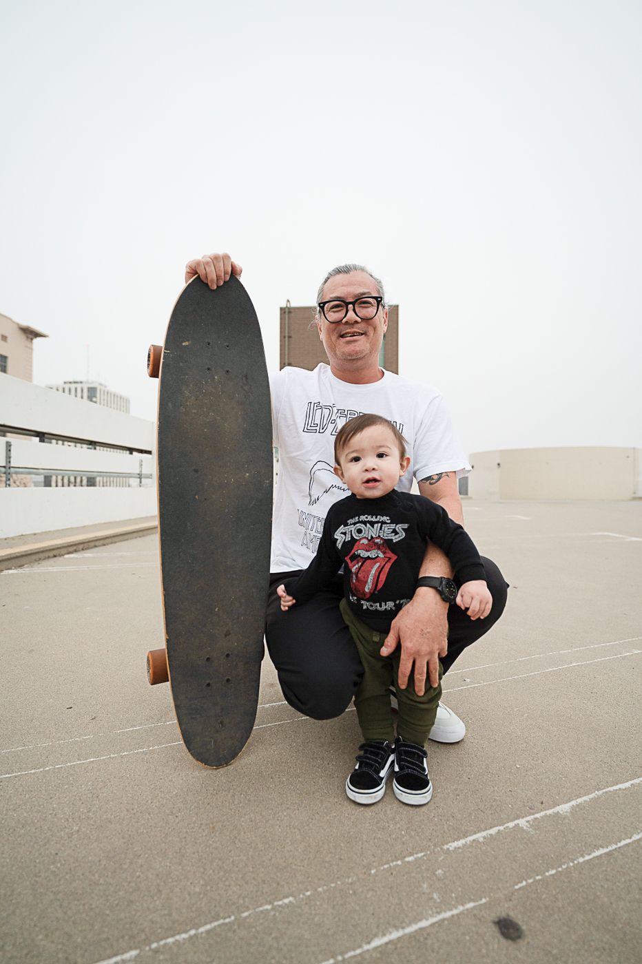 A skater poses with a longboard and young child on an urban rooftop setting.
