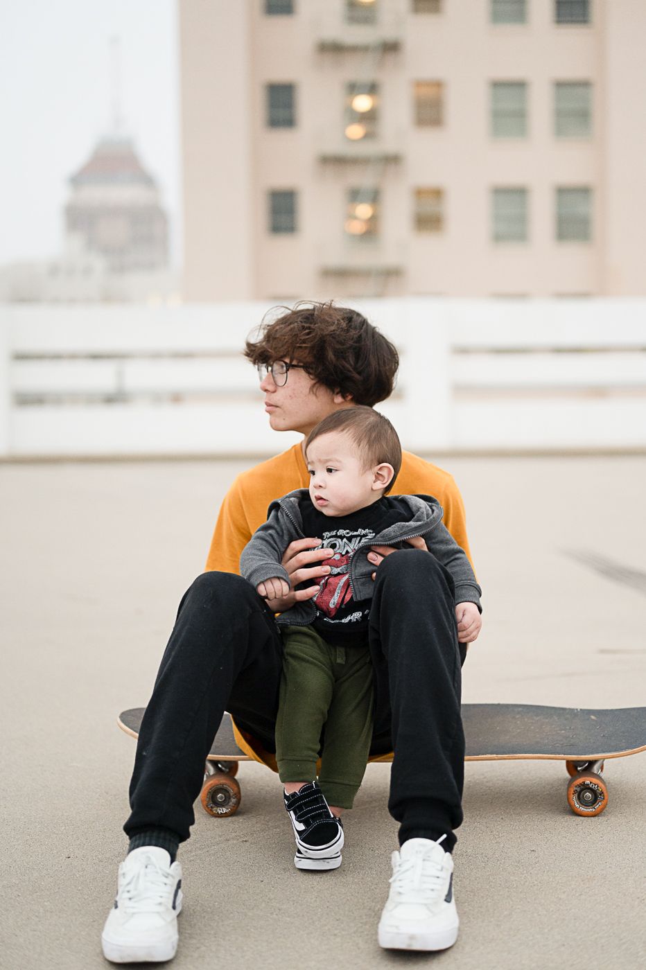 Person in orange shirt sits on skateboard with small child against city building backdrop.