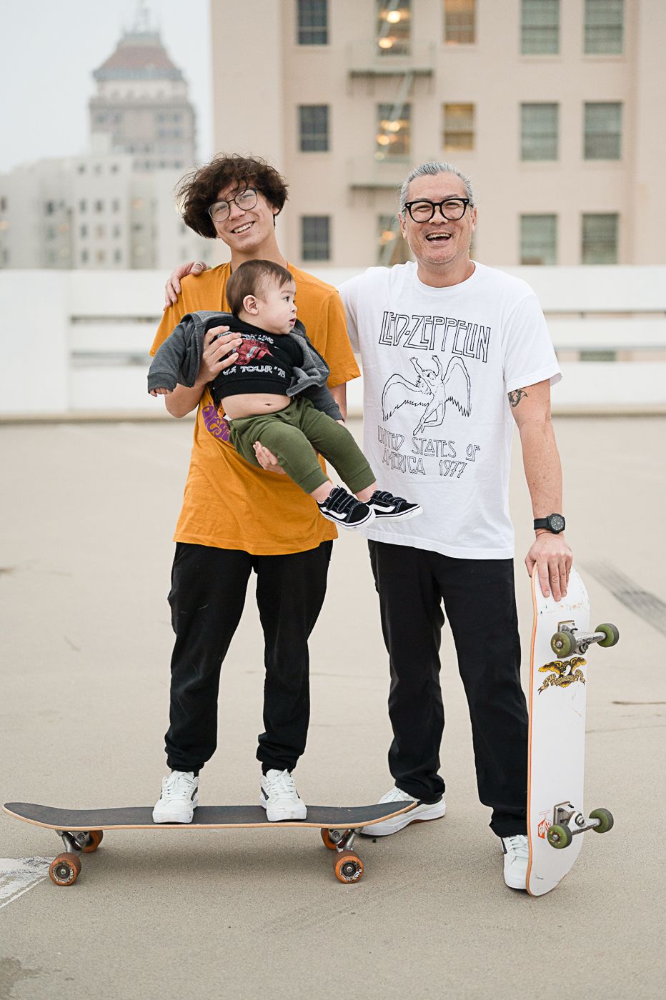 Three people pose together on a rooftop with a skateboard and city buildings in background.