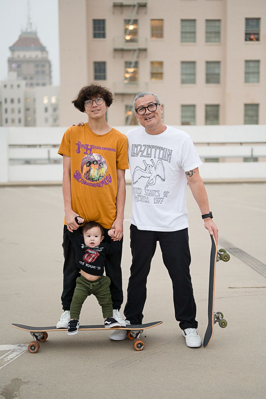 Family group poses with skateboard on urban rooftop with city architecture behind them.