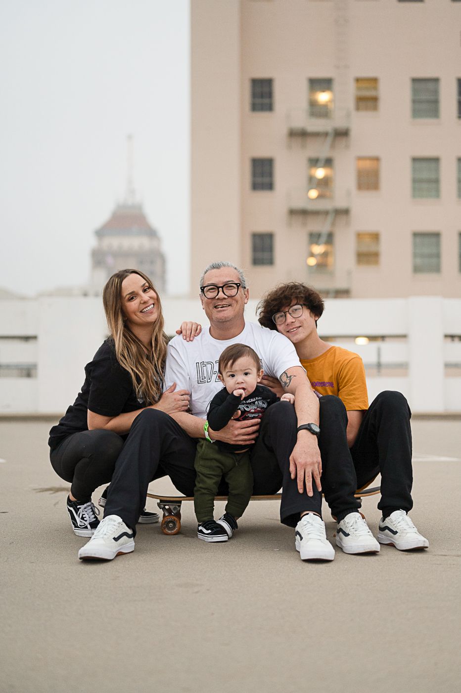 Group portrait of four people sitting together on a skateboard against city backdrop.