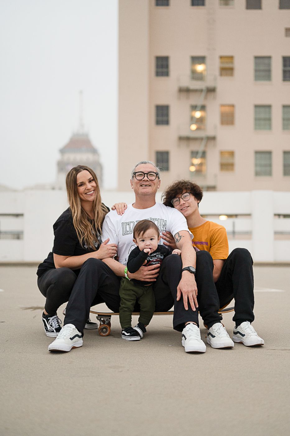 Family of four poses together on rooftop with skateboard and urban skyline.
