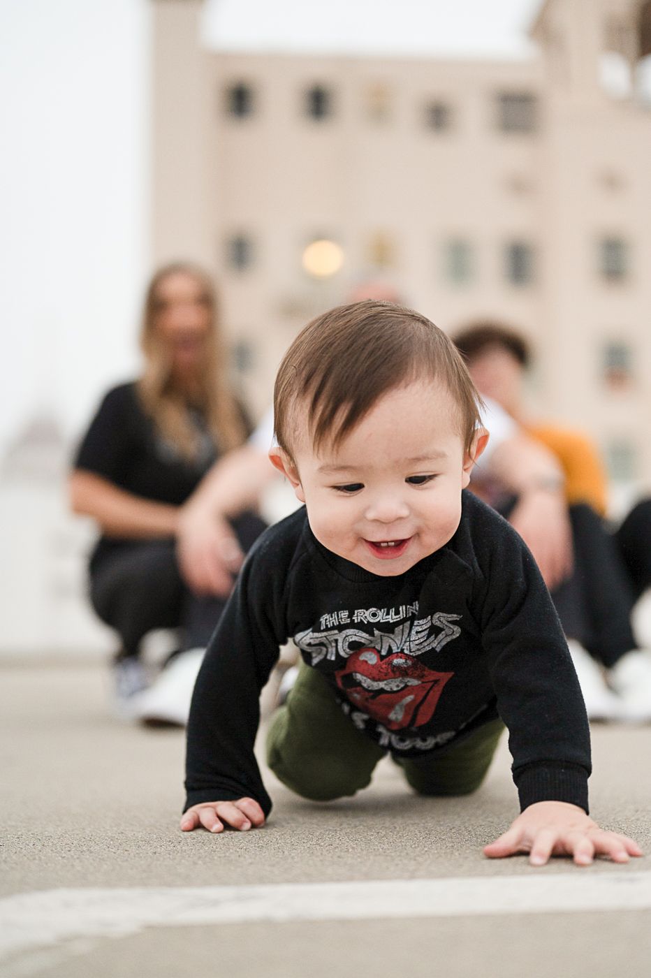 Smiling baby crawls on concrete surface wearing black sweatshirt.