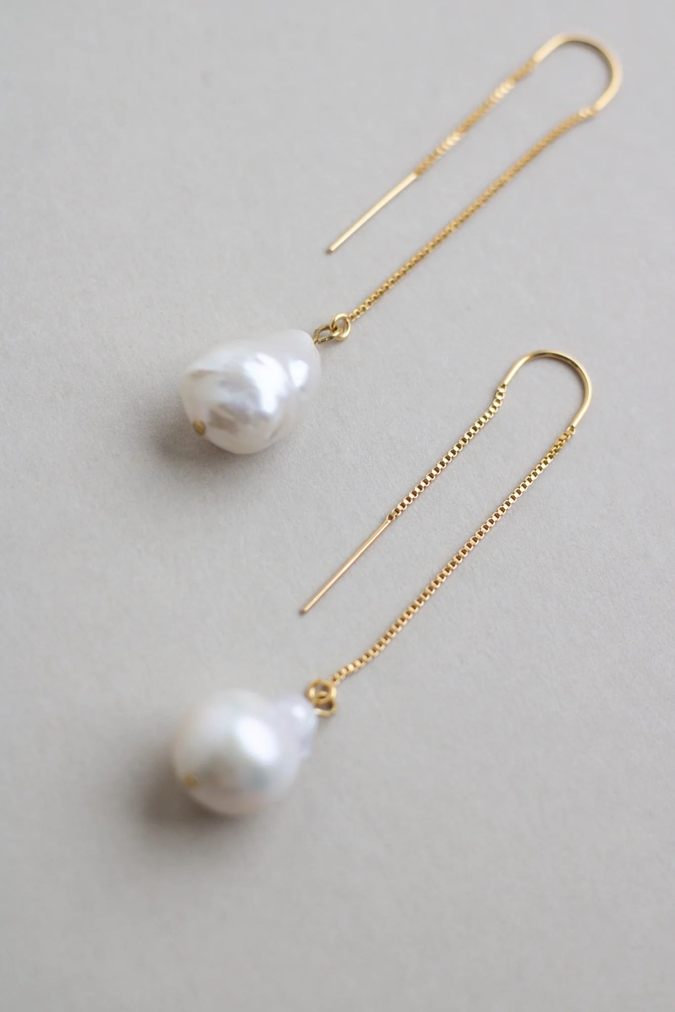 Minimalist gold threader earrings with baroque pearls on light gray background.