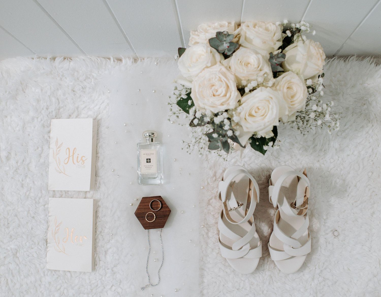 Wedding details flatlay with white roses, invitation cards, and strappy sandals on white surface.