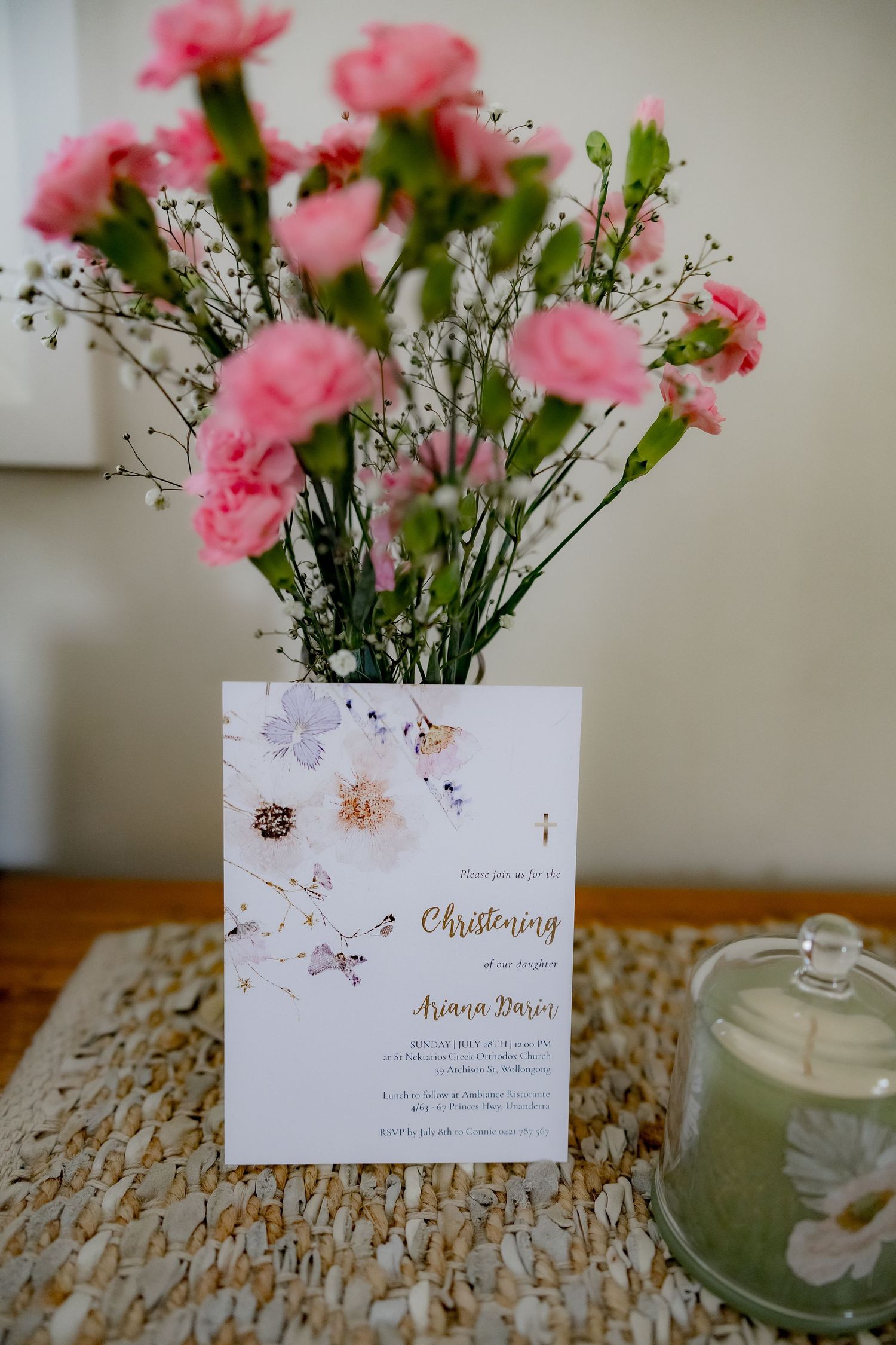 Pink carnations in a vase next to a christening invitation and green candle on a textured table surface.