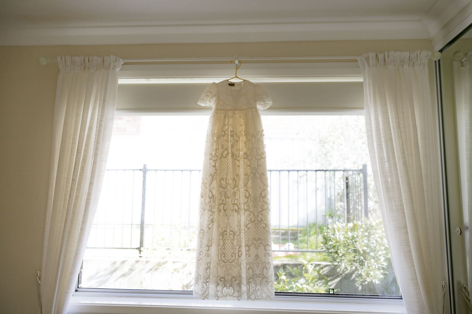 White lace dress hanging in window with sheer curtains and natural lighting.