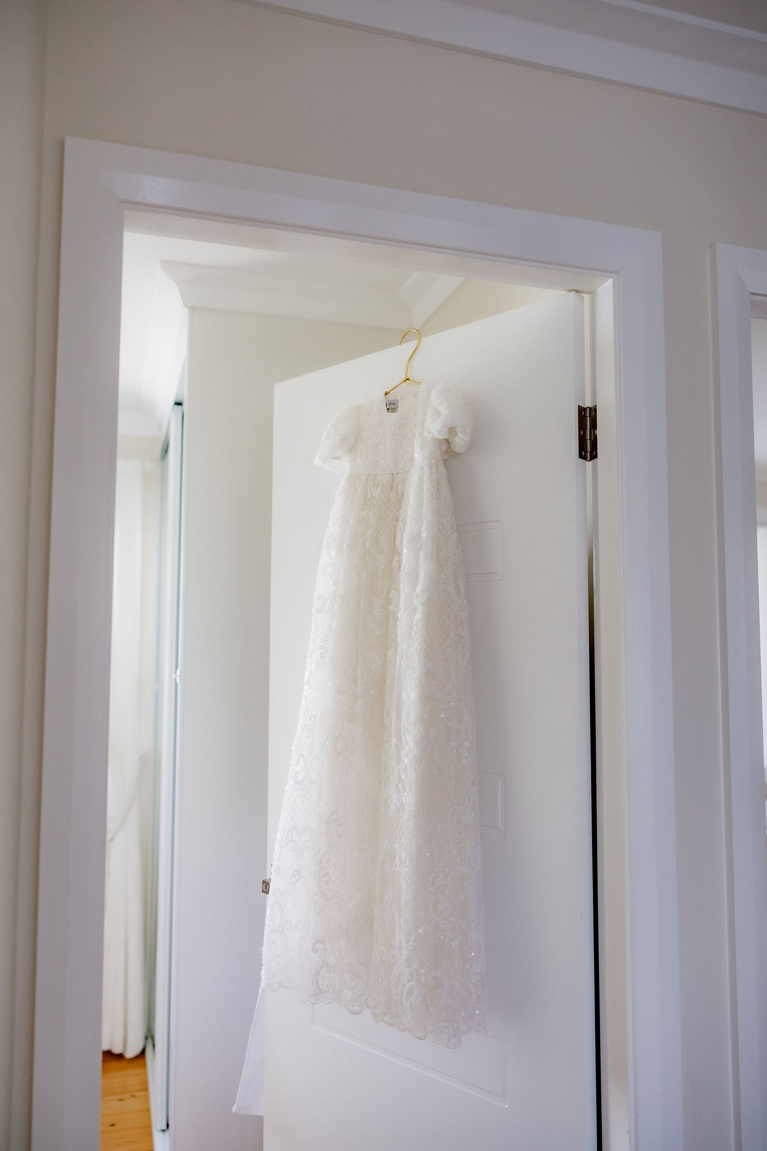 White christening gown hanging in doorframe against neutral wall.