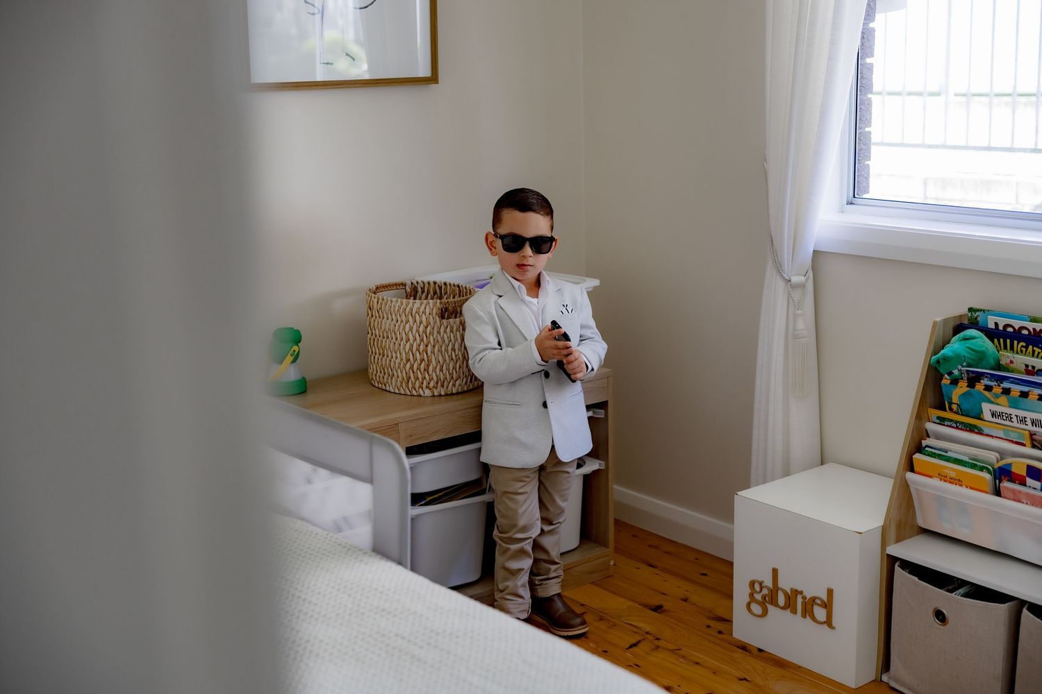 A toddler wearing sunglasses and a white outfit stands in a nursery room with toys and storage baskets.
