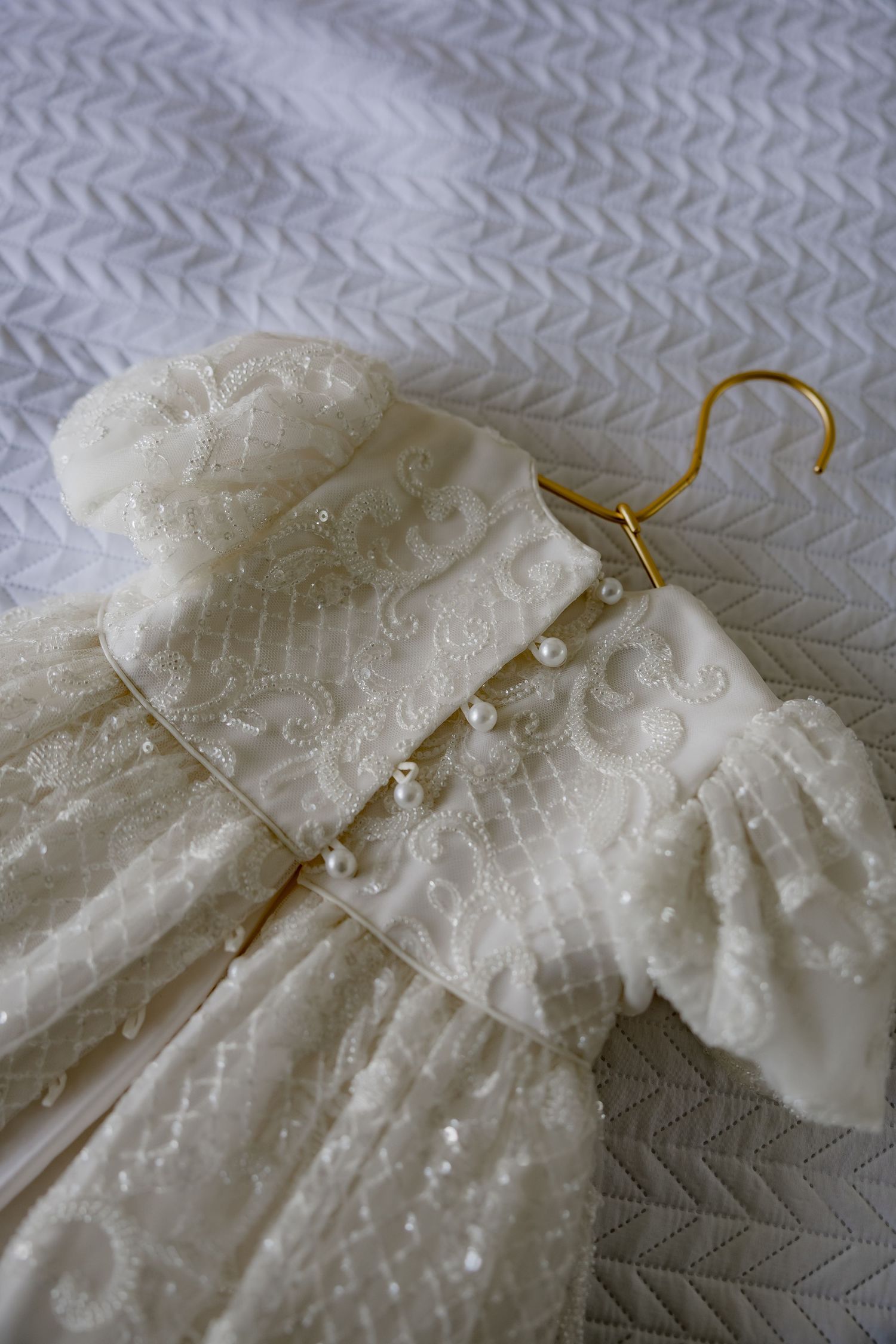 A delicate white baptismal gown with pearl details hangs on a gold hanger against a patterned fabric background.