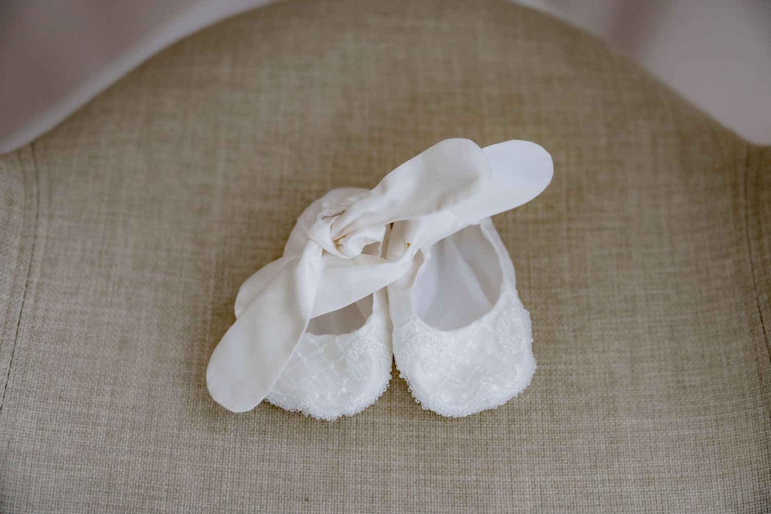 Dainty white lace baby shoes displayed on a neutral linen surface.