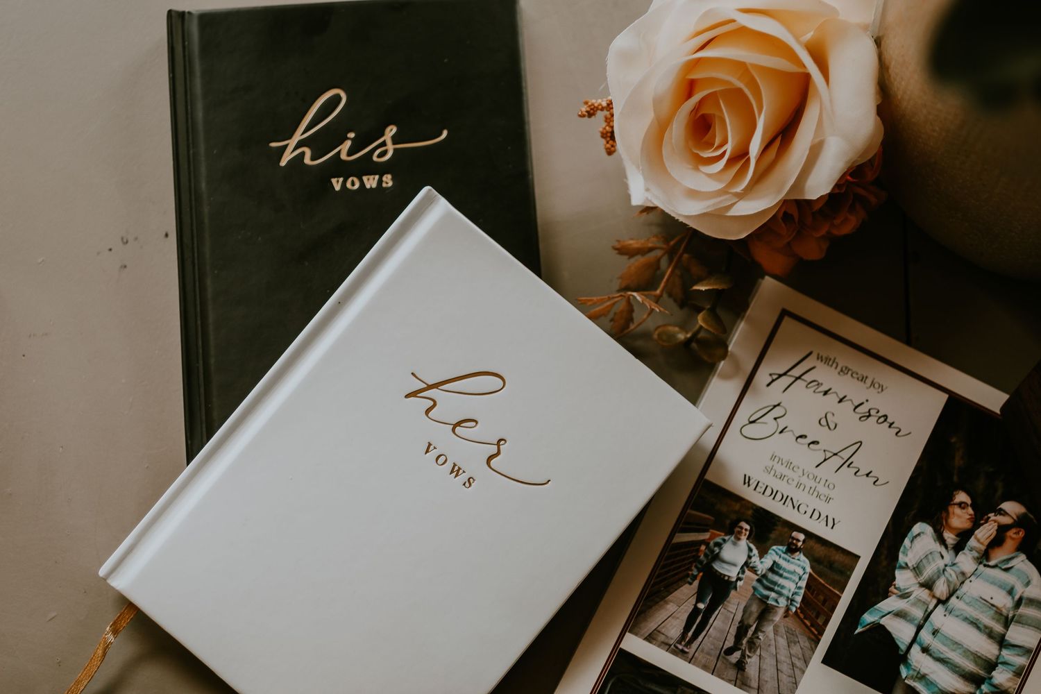 His and her vow books with gold foil lettering beside a peach rose and wedding program on a flat lay.
