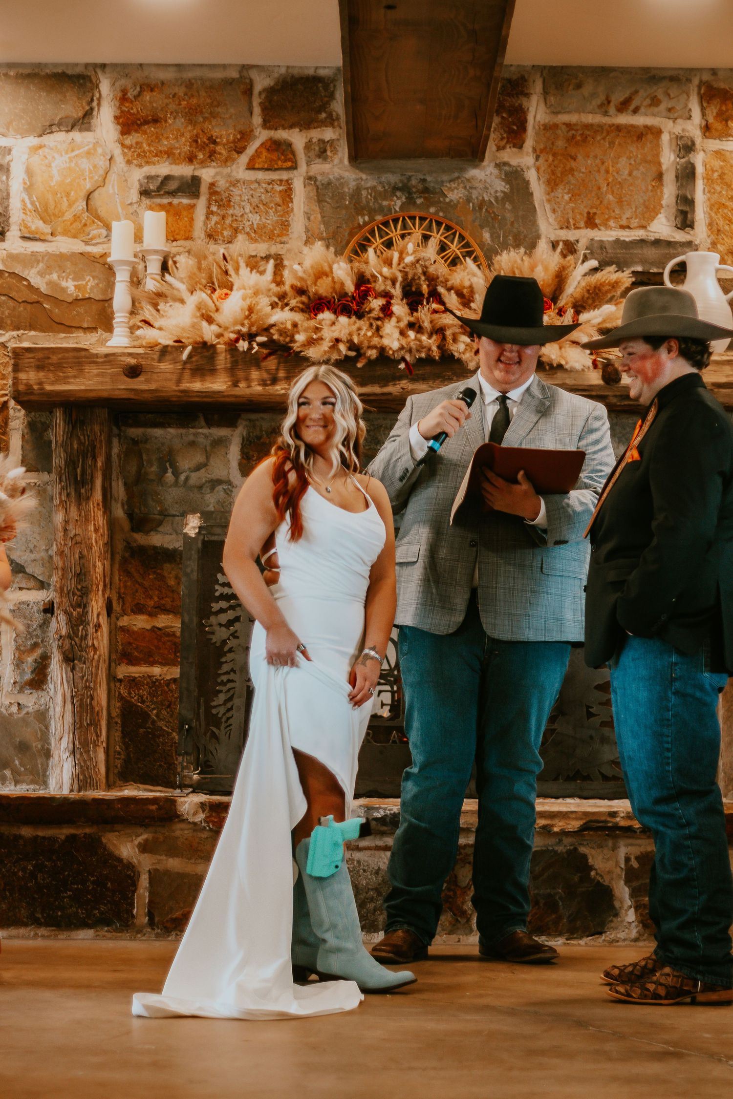 Bride in white gown and turquoise cowboy boots smiles during rustic wedding ceremony by stone fireplace.