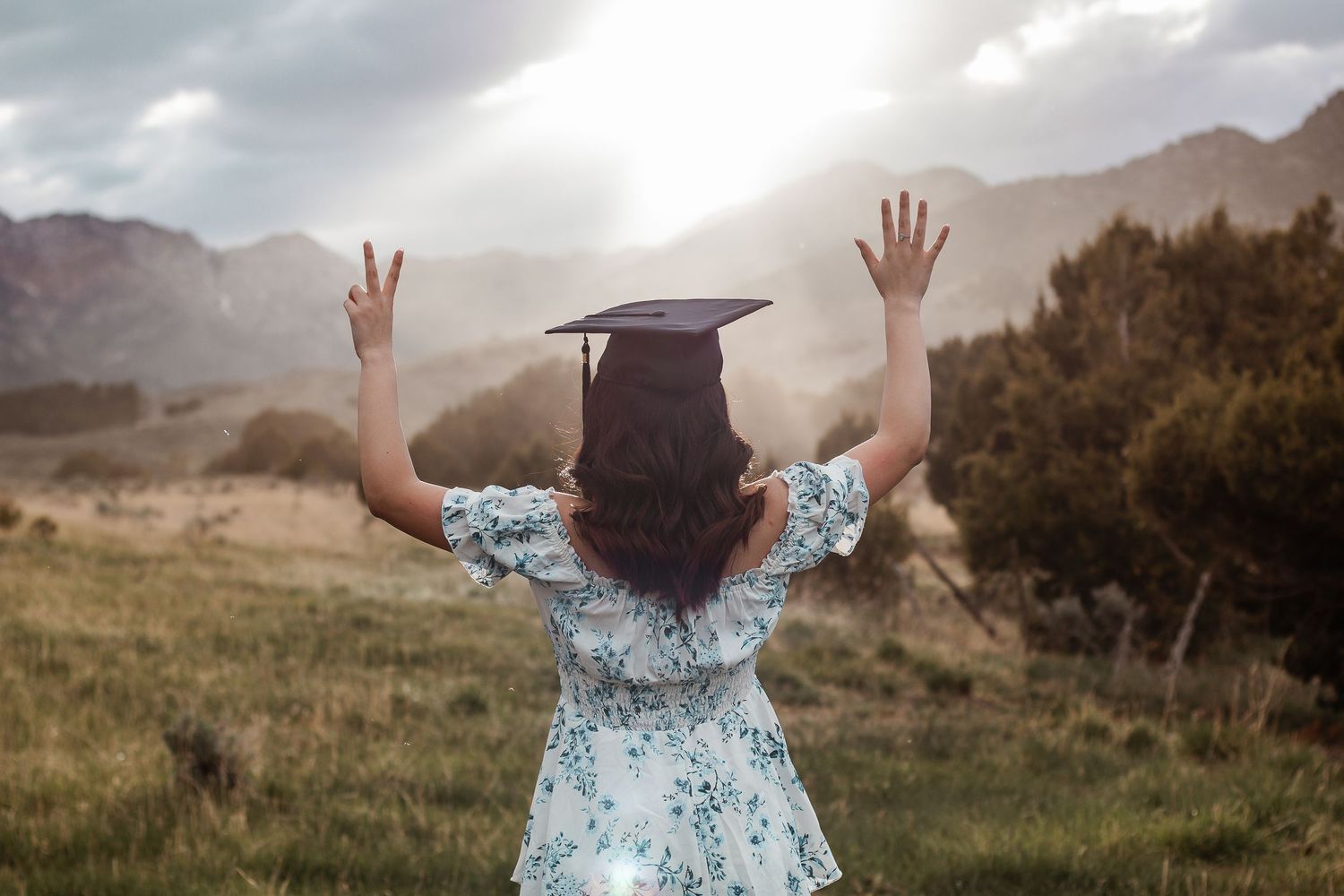 A young woman in a floral dress and graduation cap raises her hands triumphantly in a scenic mountain meadow.