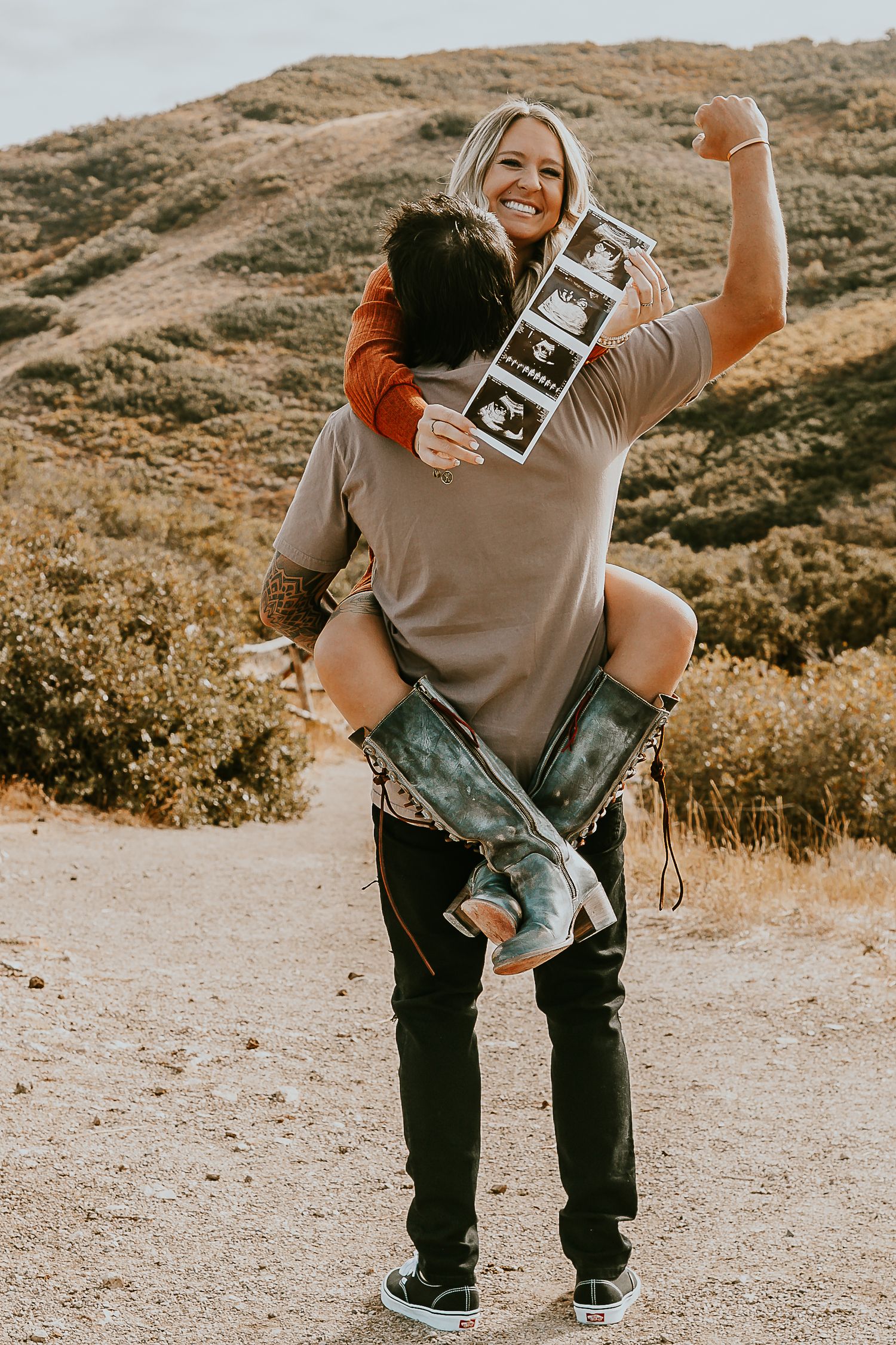 Couple celebrating pregnancy announcement outdoors, woman on man's back holding ultrasound photos, hills in background.