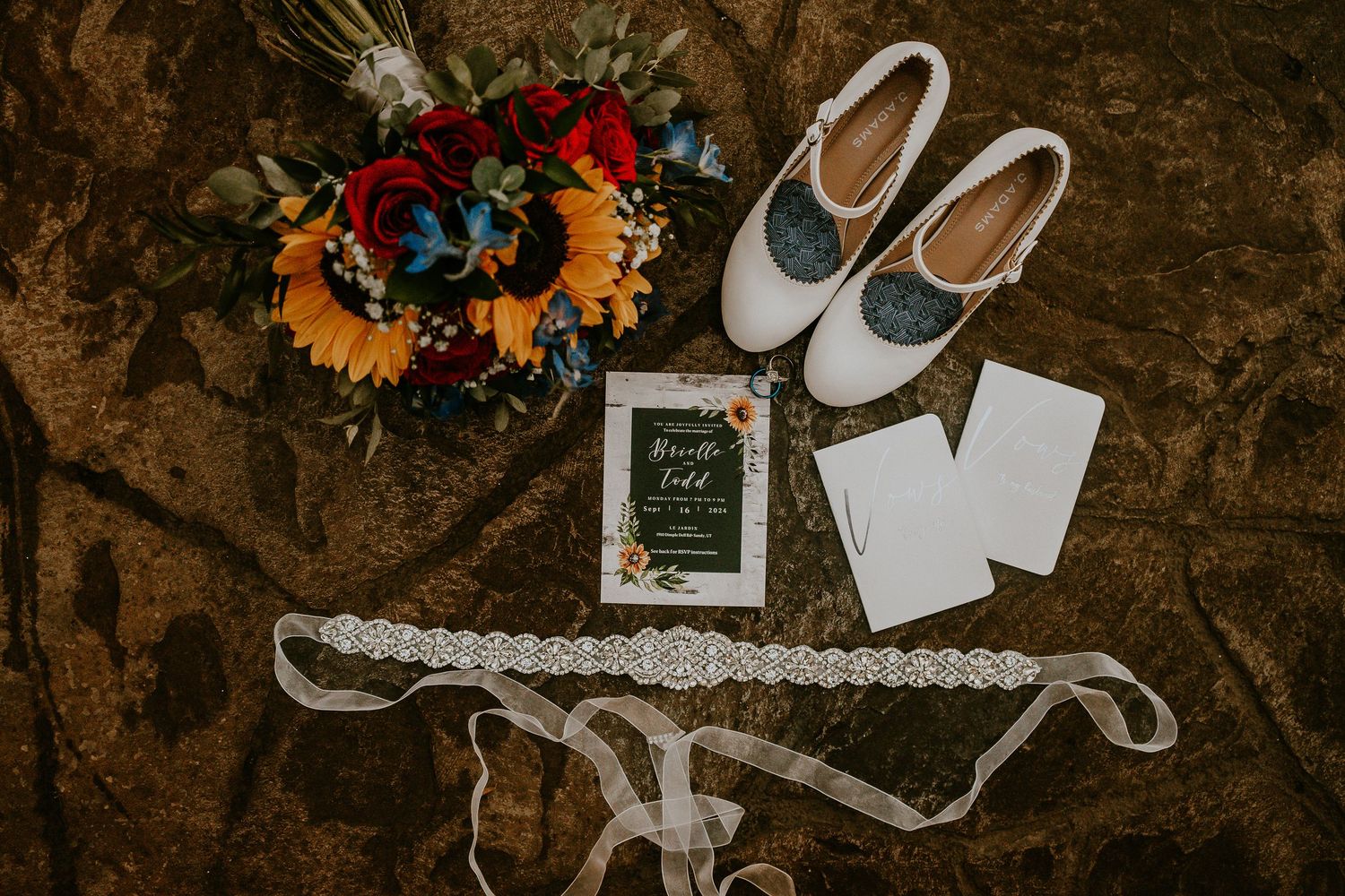 Wedding flat lay featuring sunflower bouquet, white heels, invitation suite, and lace bridal belt on rustic stone surface.