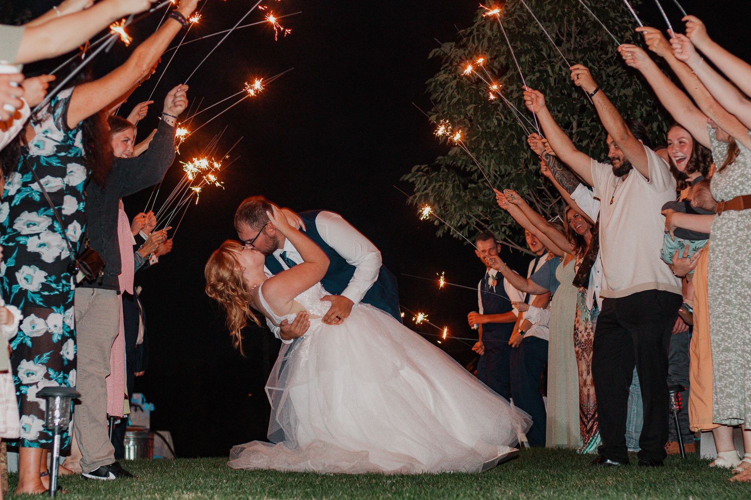 Bride and groom share a romantic dip kiss surrounded by guests holding sparklers at an outdoor wedding reception at night.