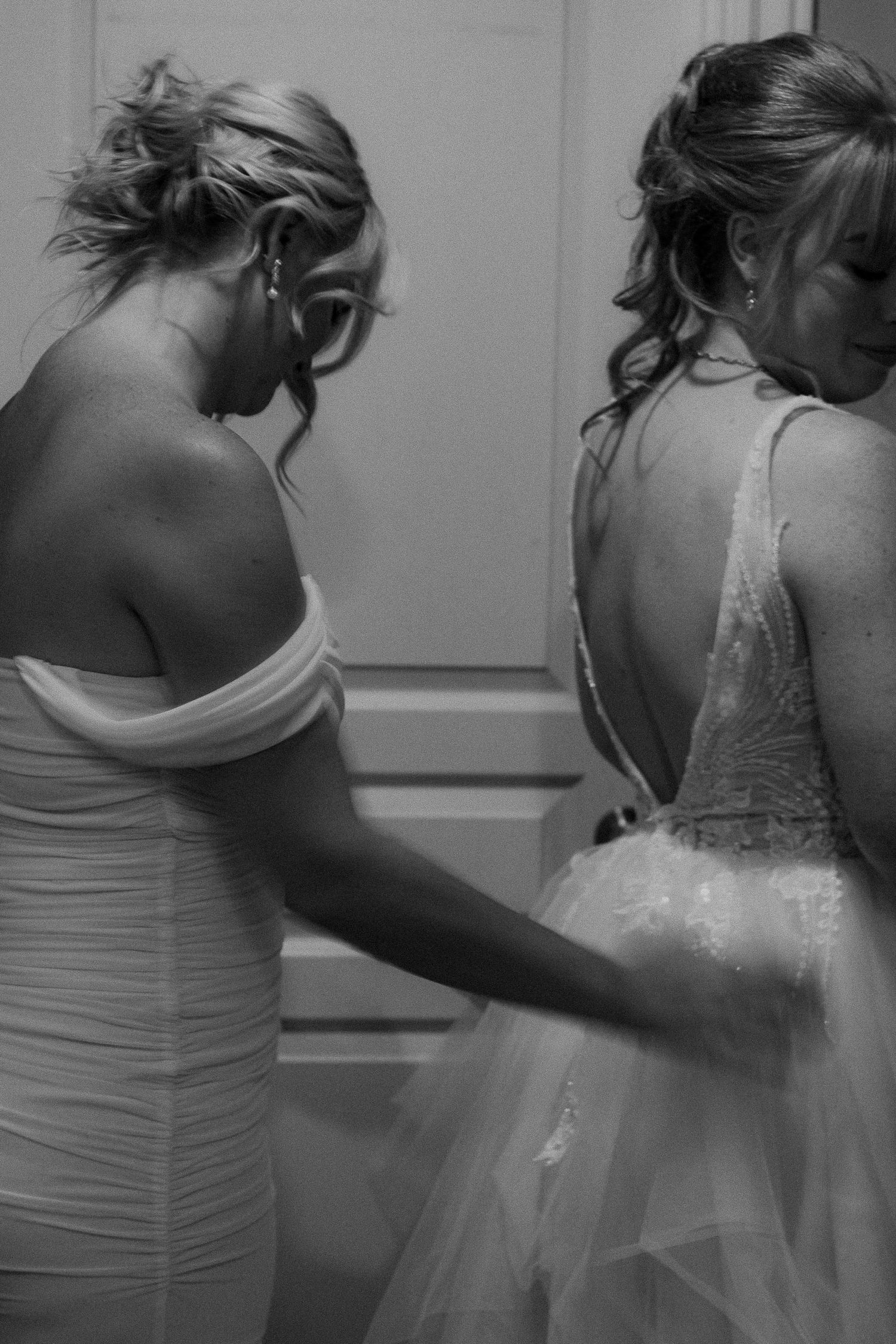 Black and white photo of a bridesmaid helping a bride with her open-back wedding gown before the ceremony.