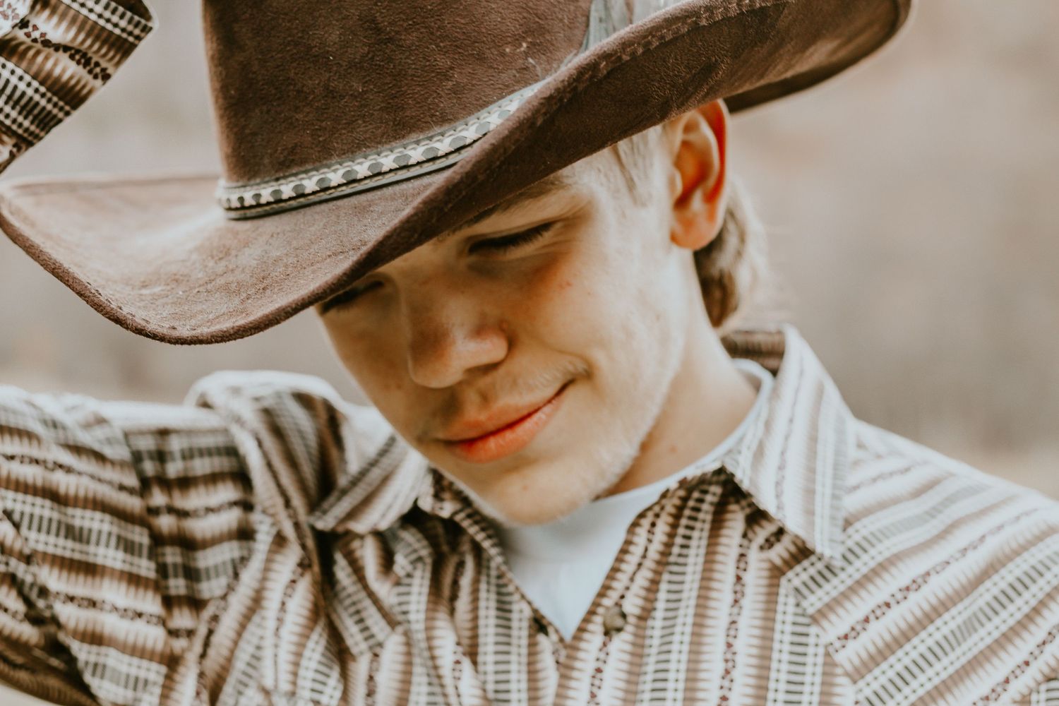 Young cowboy in a worn brown hat and striped western shirt smiling downward in a rustic outdoor setting.