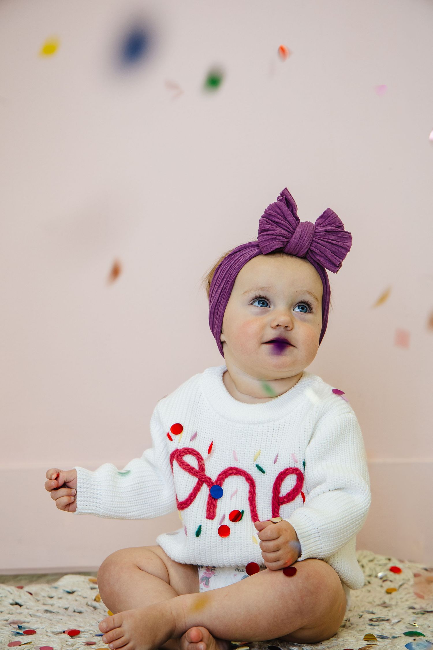 Baby girl in white 'One' sweater and purple bow headband sits among colorful confetti celebrating her first birthday.
