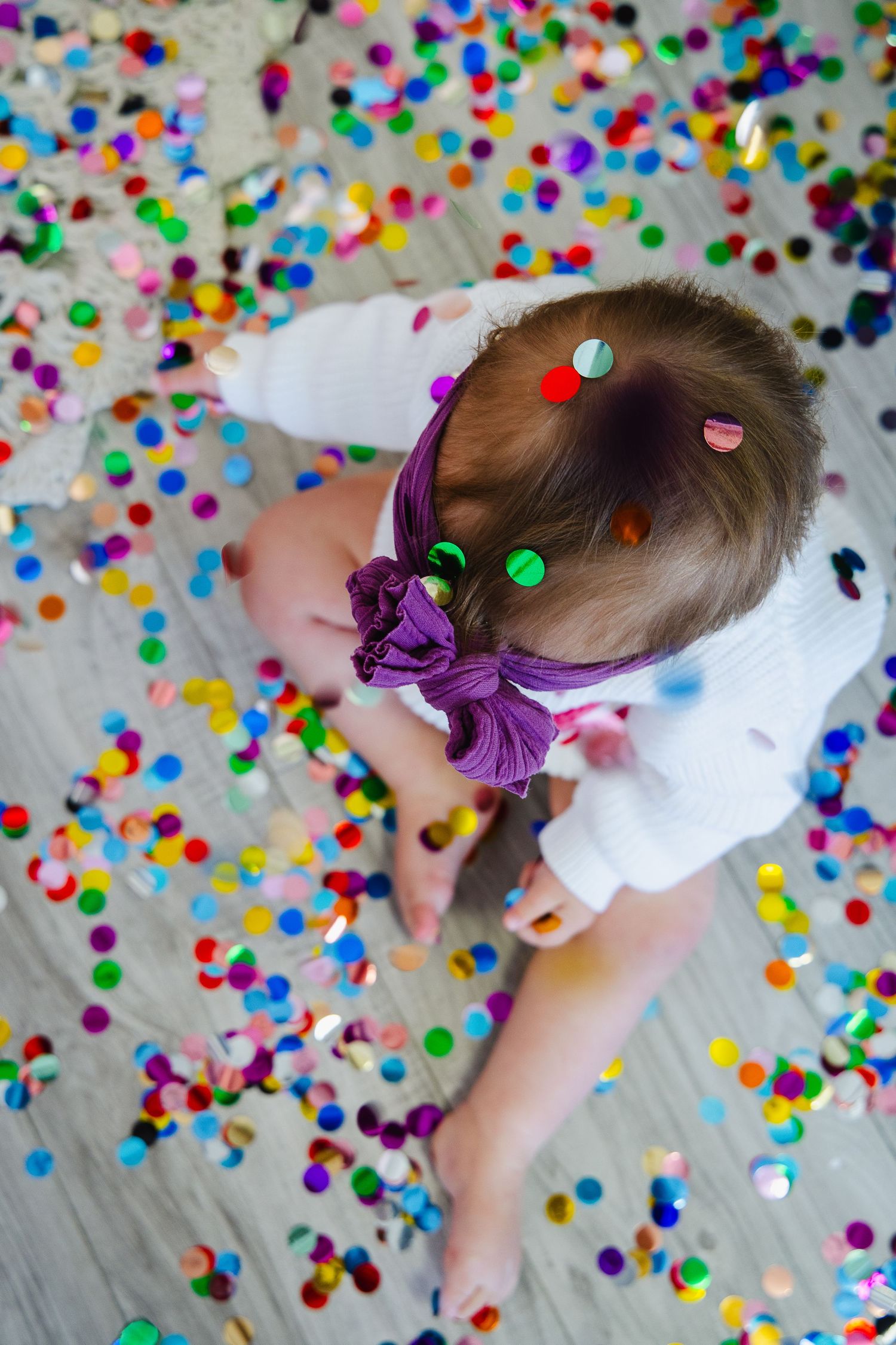 Toddler girl with purple bow headband sitting on floor covered in colorful confetti during a celebration.