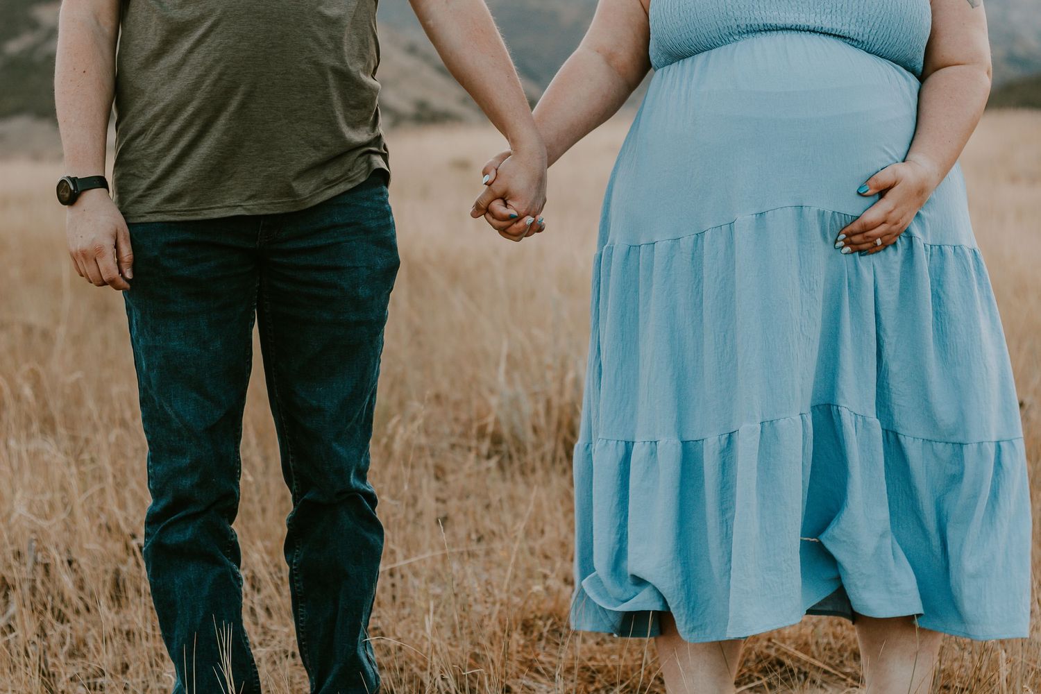 Pregnant couple holding hands in a golden field, woman in blue dress cradling baby bump.