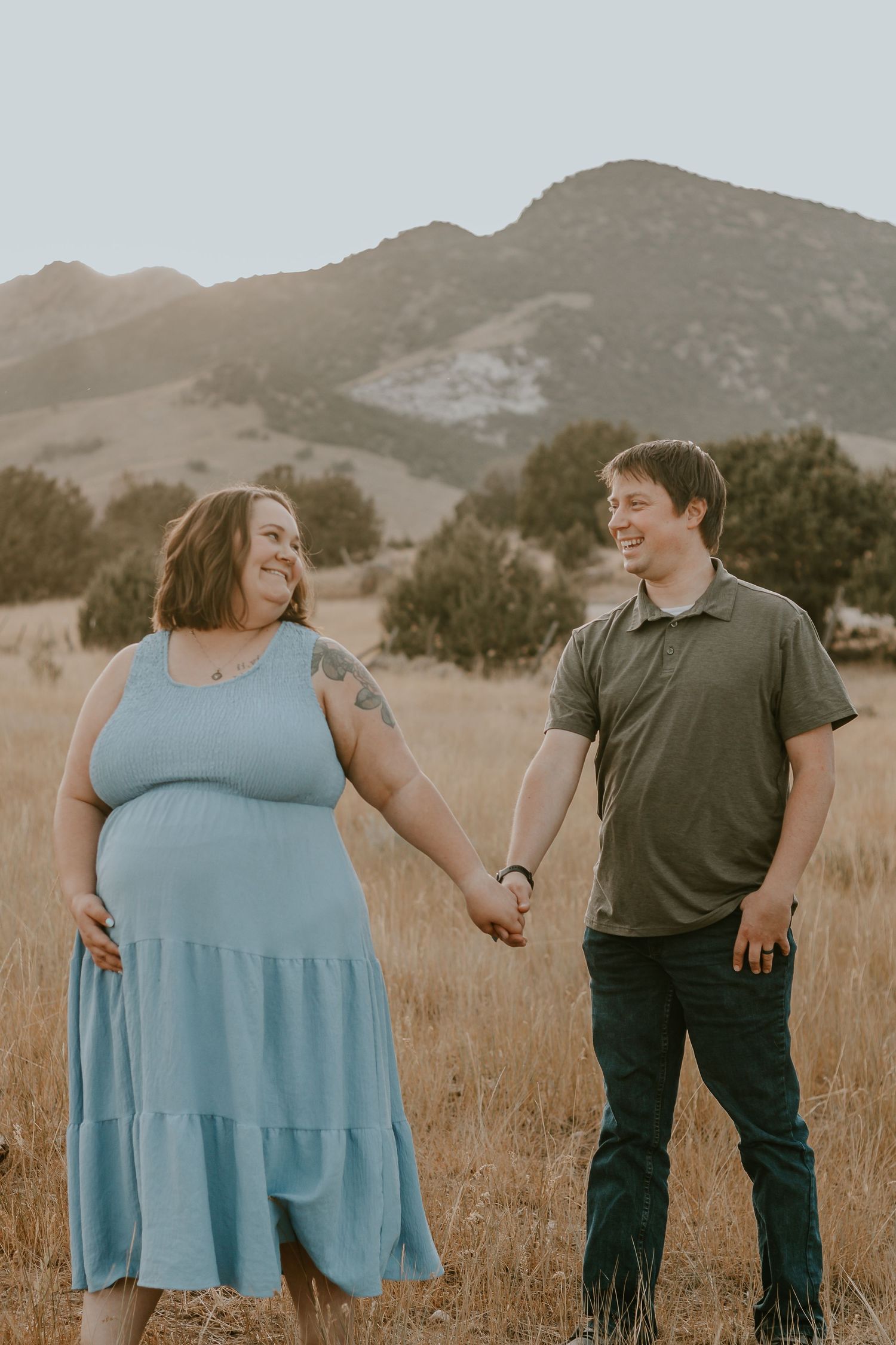 Pregnant couple holding hands smiling in golden field with mountain backdrop during maternity photo session.