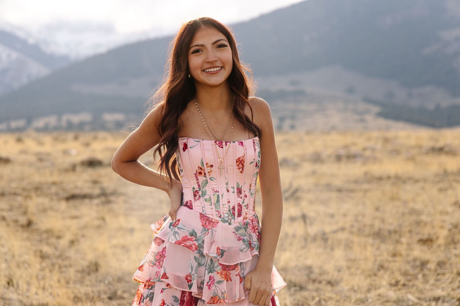 Young woman in floral pink sundress smiling outdoors with mountain landscape and golden fields in background.