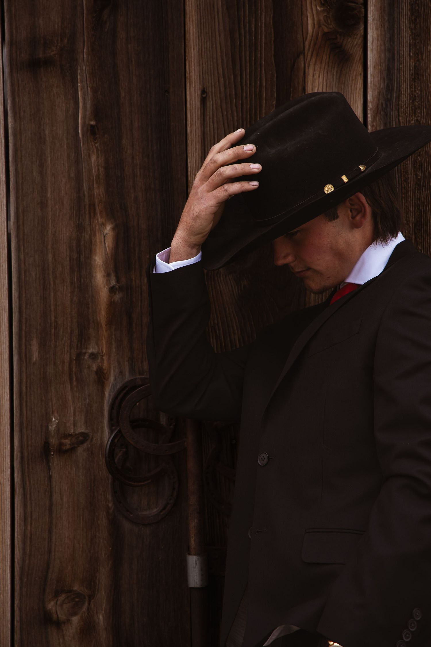 Man in black cowboy hat and suit leaning against rustic wooden wall, looking down dramatically.