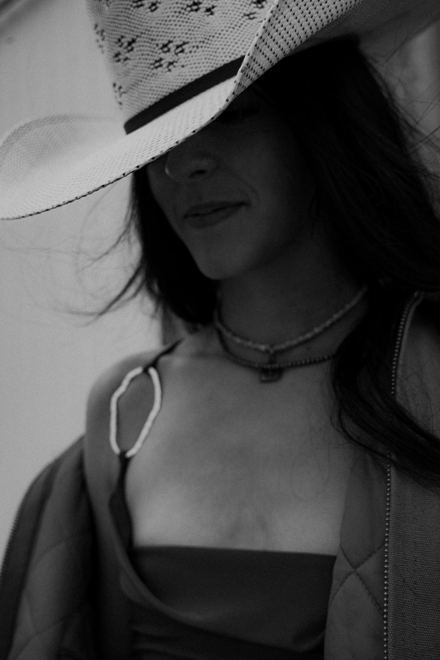Black and white portrait of a woman in a cowboy hat and layered necklaces, smiling softly.