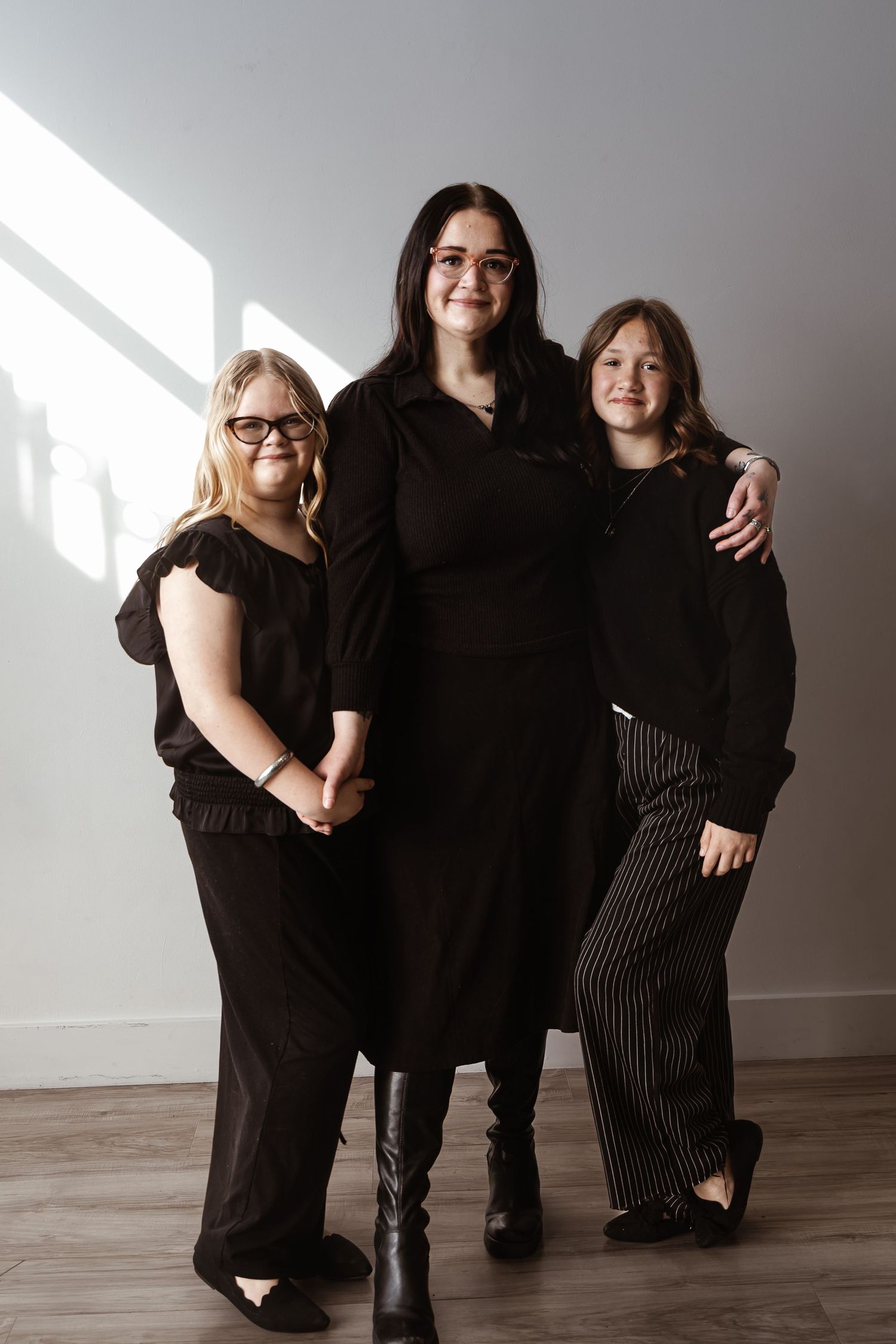 Three young women in black outfits pose together smiling in a bright studio setting.